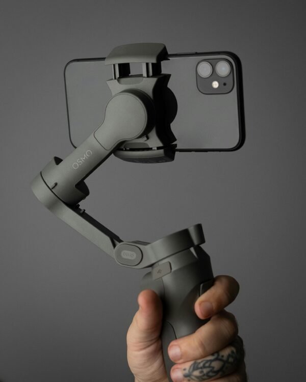 Hand holding a smartphone mounted on a stabilizer against a solid background with tattooed fingers.